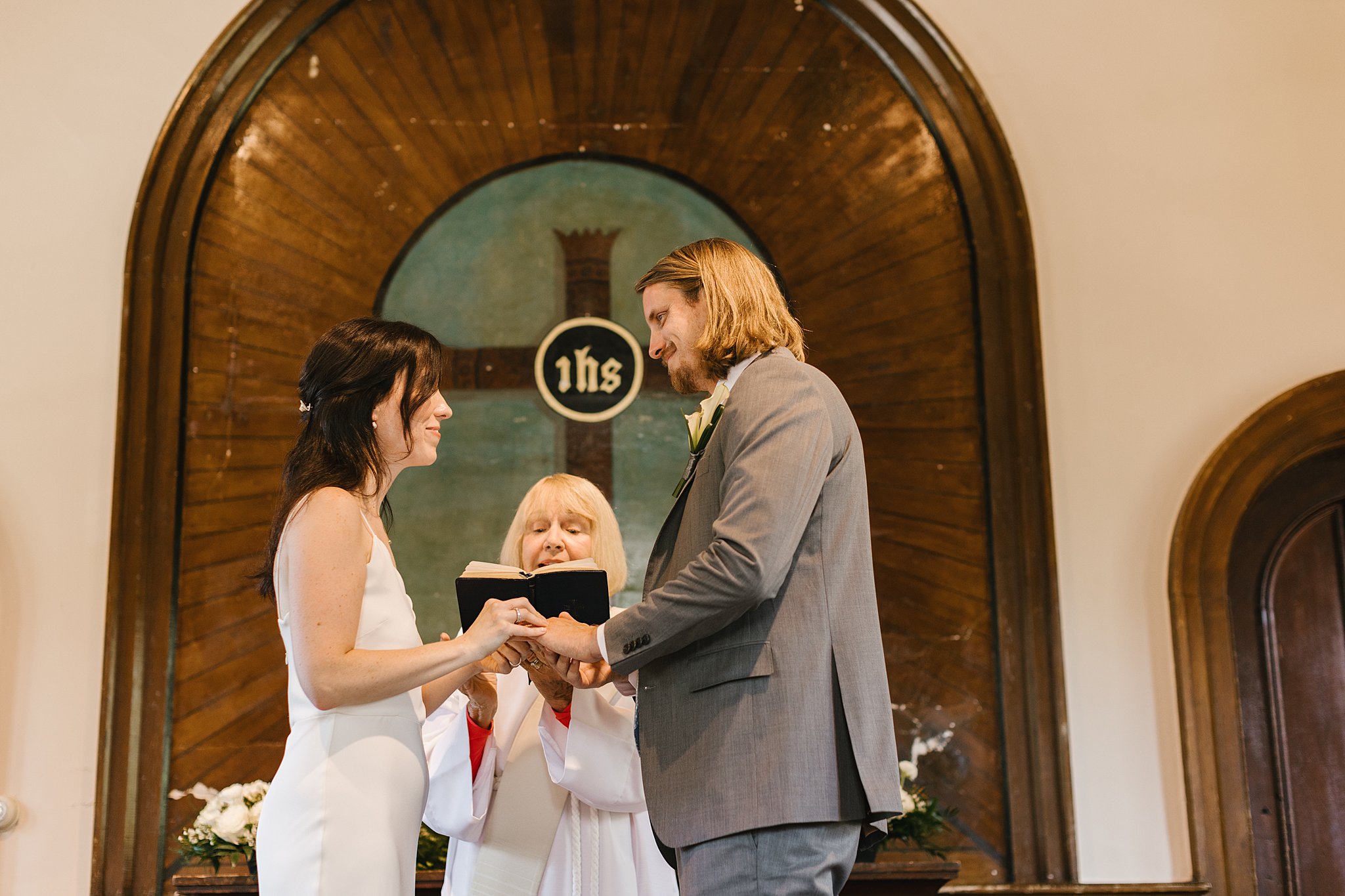 Bride and groom hold hands and exchange rings as their priest reads at the altar