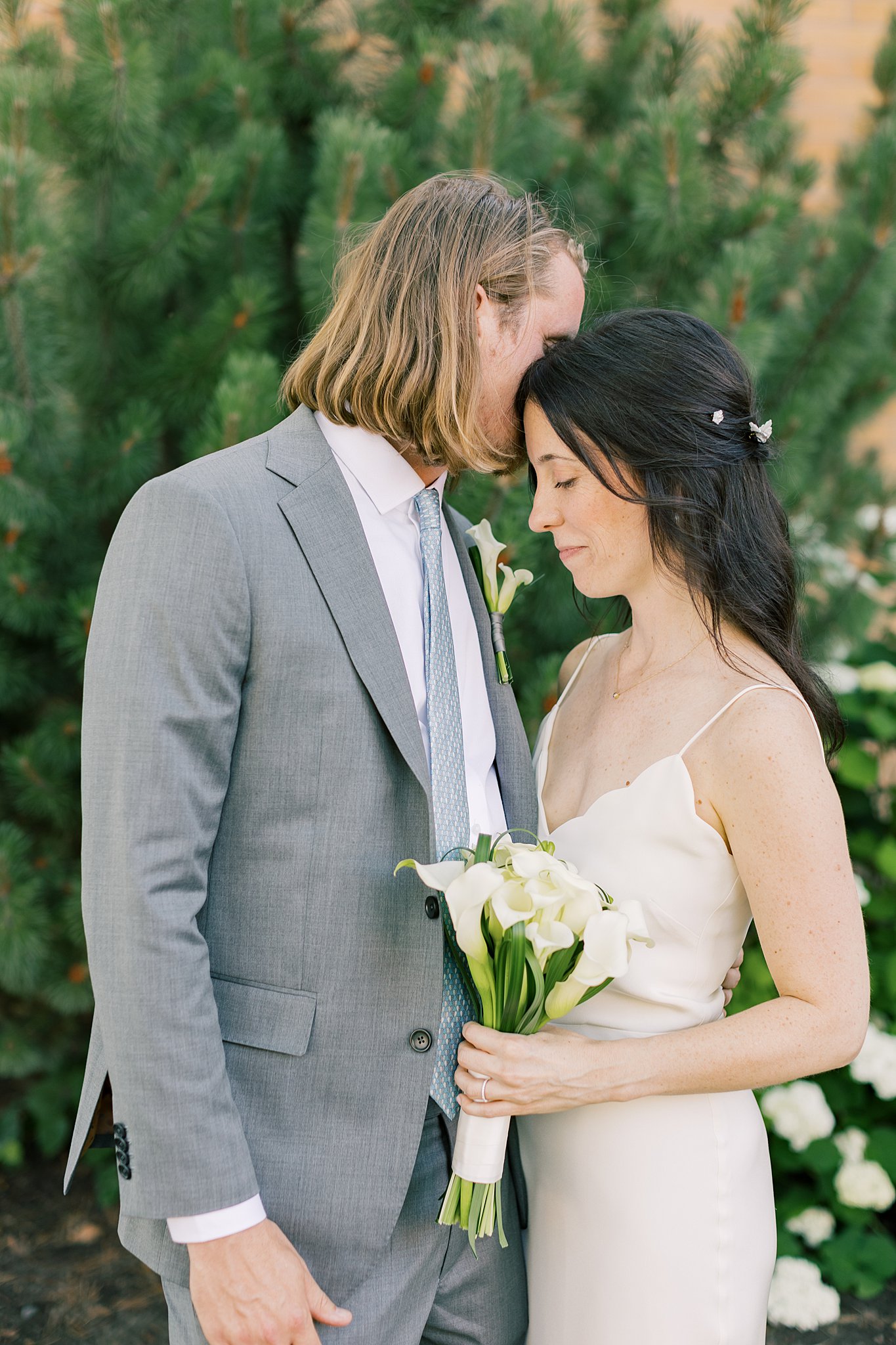 Newlyweds cuddle while standing in a garden in a grey suit and silk dress with a white bouquet at fort sherman chapel