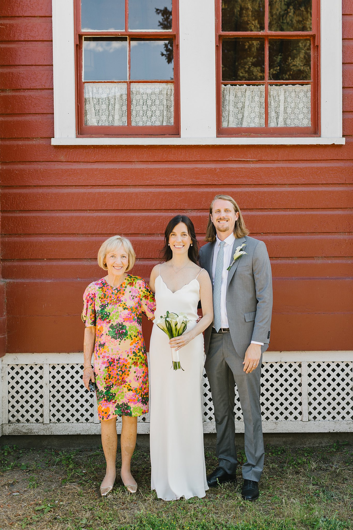 A bride and groom stand with a woman in a bright dress smiling