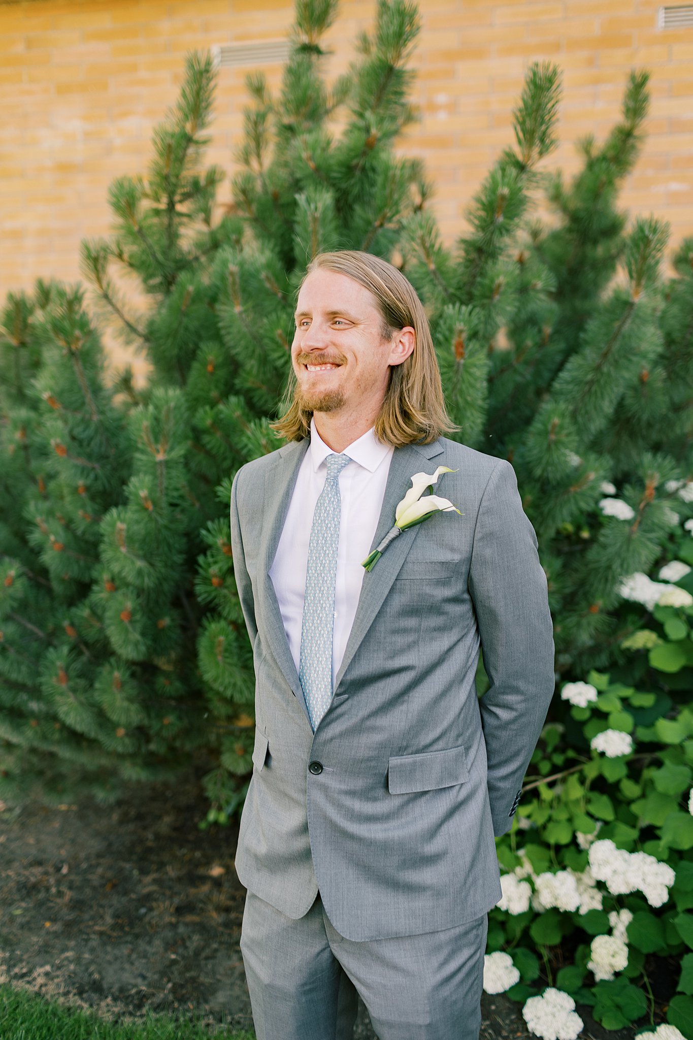 A groom smiles big in a grey suit while standing in a garden with hands behind his back at his fort sherman chapel wedding