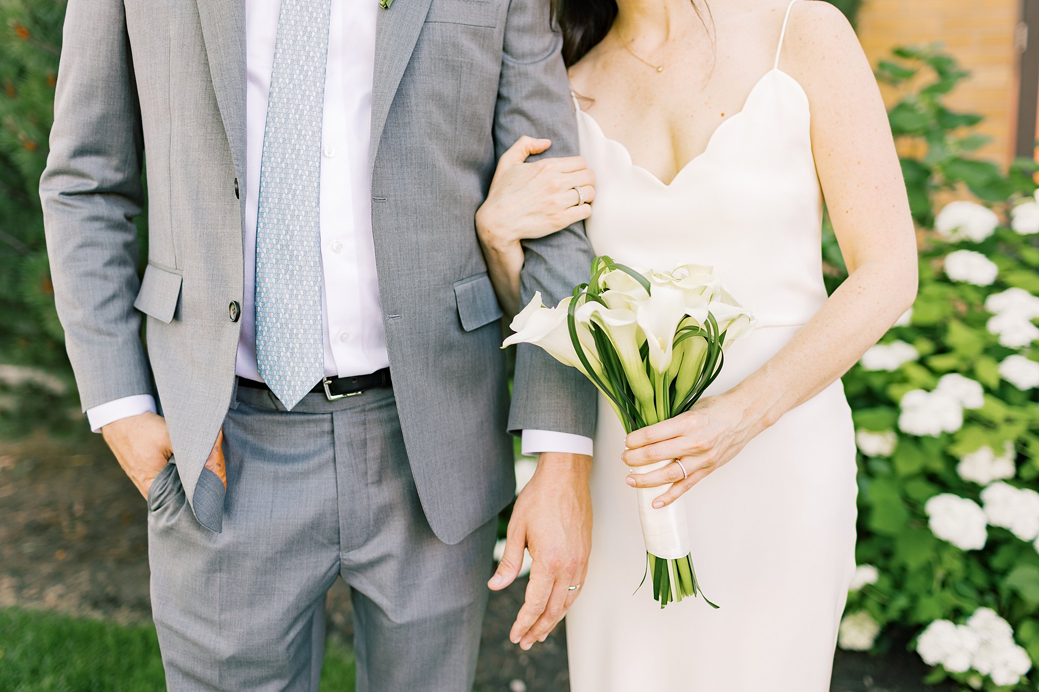 A bride and groom stand in a garden holding a white bouquet