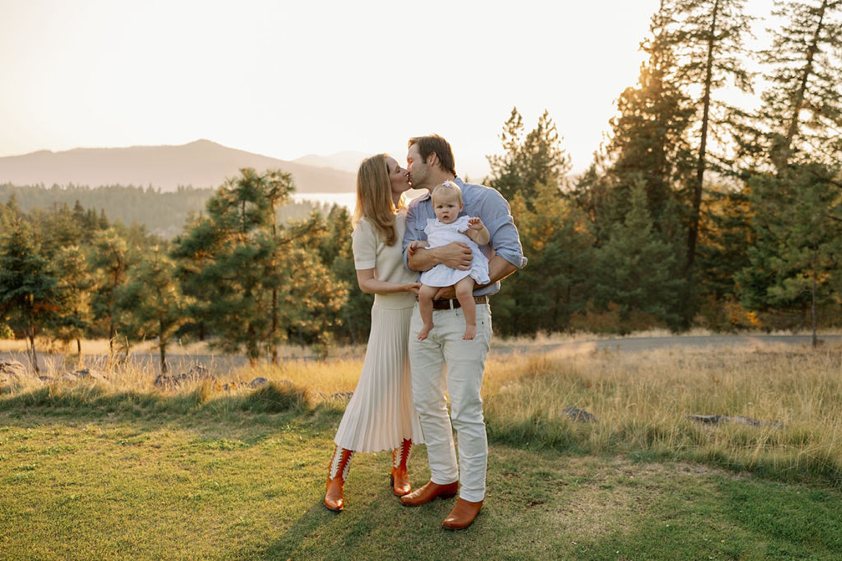 A mom and dad kiss while dad holds their young toddler daughter in blue and cream at sunset on a mountain lawn