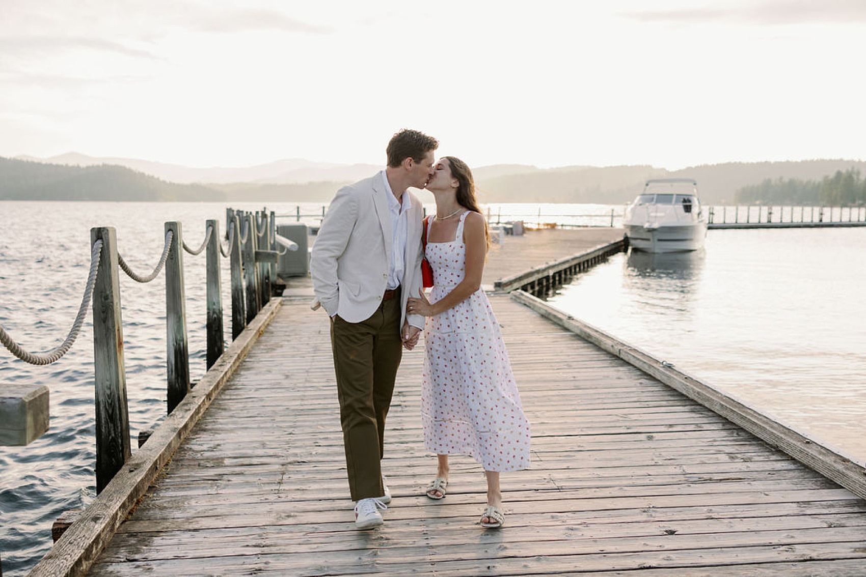 A happy newly engaged couple kiss while holding hands walking on a dock at sunset during their proposal in Coeur d’Alene